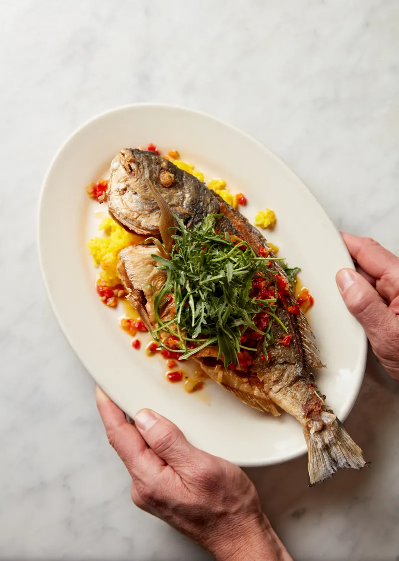 Hands presenting a whole fried fish topped with arugula and red pepper relish