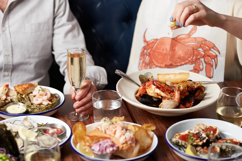 Two diners sharing cioppino, oysters, and a lobster roll with champagne